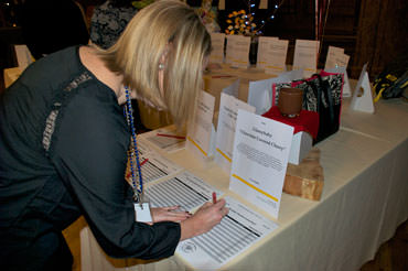 A woman fills in her bid on a ReadySetAuction bid sheet while glancing at a display sheet that describes the auction item.