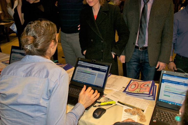 A woman running  a ReadySetAuction check-in station on a laptop talks to the next guests in line.